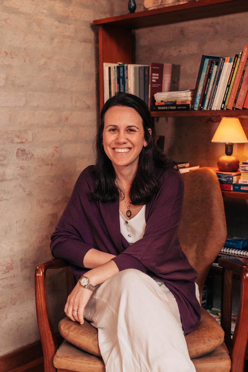 Carla Latini sorrindo sentada em uma poltrona acolhedora, com abajur e estante de livros ao fundo